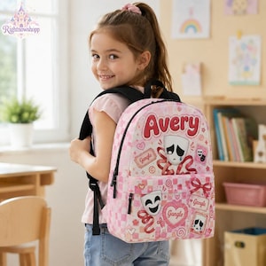 Puede incluir: Una mochila a cuadros rosas y blancos con el nombre Avery en letras rojas, junto con ilustraciones de máscaras de teatro, cintas y corazones. La mochila incluye correas y cremalleras negras, con el texto Gangle impreso en etiquetas.