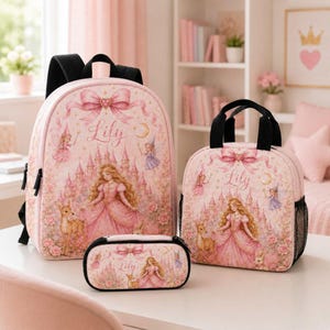 May include: A pink backpack, lunch bag, and pencil case set featuring a printed illustration of a princess in a gown, a castle, a deer, and a rabbit. Each item displays a large bow at the top and the name Lily in cursive script text.