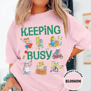 May include: A light pink t-shirt featuring the green text KEEPING BUSY and various illustrations of anthropomorphic animals engaged in different activities, including a cat with a bucket, a bear at a desk, and a character riding a red motorcycle.
