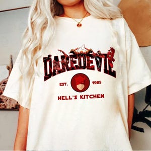 May include: Cream-colored t-shirt featuring the text "DAREDEVIL" in a red and black design, with a graphic of the character and the words "EST. 1985 HELL'S KITCHEN" in red. The shirt has short sleeves.
