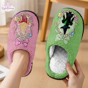 May include: A pair of fuzzy slippers, one pink and one green, each featuring a decorative oval frame graphic. The pink slipper displays a blonde figure in a crown, while the green slipper shows a dark-clad figure with a pointed hat and bow accents.