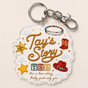 May include: A clear acrylic keychain featuring the text Tay's Story in a rope-like font, a sheriff badge with Infinity and Beyond, red cowboy boots, a red hat, colorful beads with You've got a friend in me, blocks with TS5, and clouds.