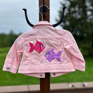Puede incluir: Una chaqueta vaquera de color rosa claro con cuello, mangas largas con puños abotonados y cierre frontal de botones. La parte trasera de la chaqueta está decorada con dos parches bordados de peces, uno rosa brillante y otro morado, ambos con ojos blancos.