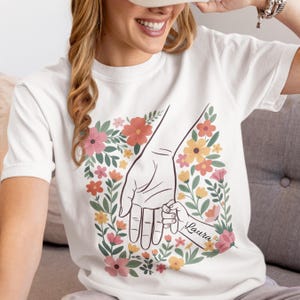 Puede incluir: Camiseta blanca con un diseño floral que representa la mano de un padre sosteniendo la mano de un niño. El diseño incluye el nombre "Laura" en la mano del niño, rodeado de flores coloridas en tonos rosa, naranja y amarillo.