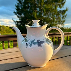 May include: A white porcelain teapot featuring a curved handle, a tapered spout, and a matching lid with a crescent-shaped finial. The body is decorated with a botanical pattern of dark blue and green maple leaves and small black berries.