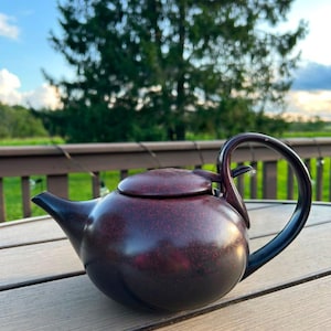 Puede incluir: Una tetera de color marrón rojizo oscuro con un acabado moteado, que presenta un cuerpo redondeado, un pico corto y un asa curva que se extiende hacia arriba y se conecta a la tapa. La tetera tiene una textura suave y mate con diseño circular.