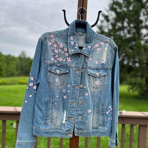 May include: A medium-wash blue denim jacket featuring hand-painted pink cherry blossom branches across the front and sleeves. The jacket includes a classic collar, button-down front, two chest pockets with button flaps, and distressed detailing.
