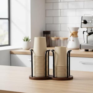 May include: A double cup holder made of dark wood and black metal, holding stacks of beige paper cups. The holder sits on a light wood surface, with a white brick wall and a window in the background. A coffee machine is visible.