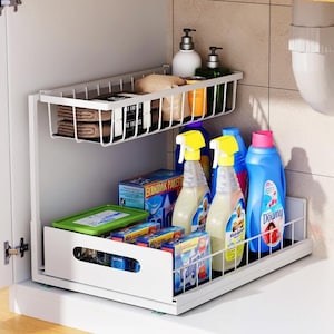 Puede incluir: Un organizador blanco de dos niveles debajo del fregadero, lleno de productos de limpieza. El estante superior contiene toallas y jabón, mientras que el estante inferior, que se desliza hacia afuera, contiene botellas de spray, detergente y cajas de productos de limpieza. El organizador está dentro de un armario blanco.