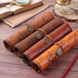 May include: Four rolled leather map journals in shades of brown and orange, each secured with a leather strap and a metal charm. The journals feature a compass rose design and a world map.