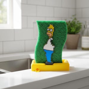 Puede incluir: Una esponja de cocina verde con Homer Simpson y un soporte amarillo. La esponja presenta una imagen de dibujos animados de Homer Simpson en amarillo, blanco y azul. La esponja se coloca en un soporte rectangular amarillo brillante, diseñado para un fregadero de cocina.
