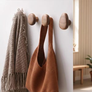 Könnte beinhalten: Drei hölzerne Wandhaken mit einer braunen Strick-Einkaufstasche und einer Fransen-Decke. Die Haken sind an einer weißen Wand montiert und bieten eine minimalistische Aufbewahrungslösung.