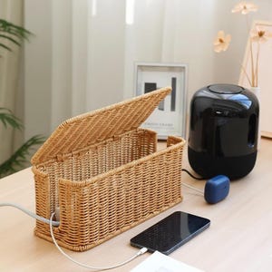 May include: A woven wicker cable organizer with an open lid. A black smartphone is charging with a white cable. A black and blue speaker and blue wireless earbuds are also visible.