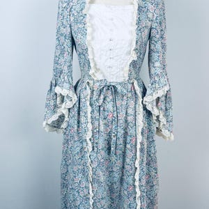 May include: A vintage-style floral print dress in shades of blue, pink, and white. The dress features long sleeves with ruffled cuffs, a white bodice with button details, and a matching lace trim. The dress is displayed on a mannequin.
