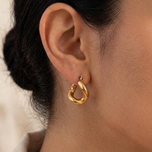May include: A close-up of a gold-colored hoop earring. The earring has a thick, rounded design and is worn on the ear. The earring is shiny and reflects light. The background is blurred, focusing attention on the jewelry.