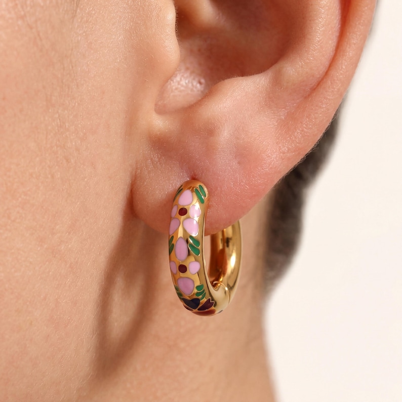 A close-up of an ear wearing a gold hoop earring with pink, green, and blue enamel floral designs.
