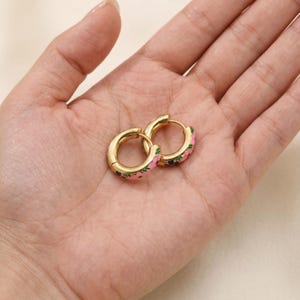 Two gold hoop earrings with pink, green, and blue enamel floral designs are held in the palm of a hand.