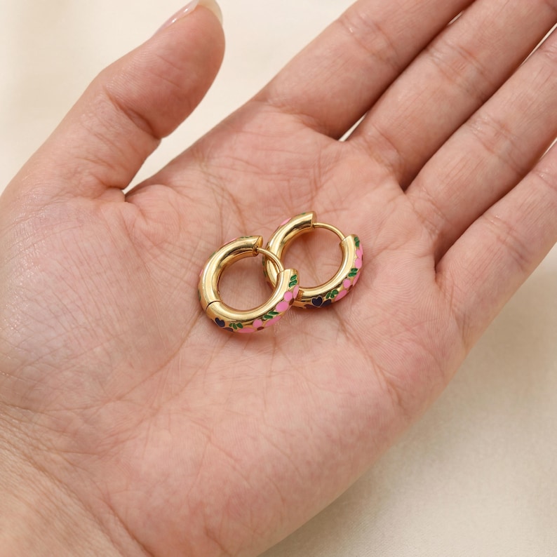 Two gold hoop earrings with pink, green, and blue enamel floral designs are held in the palm of a hand.