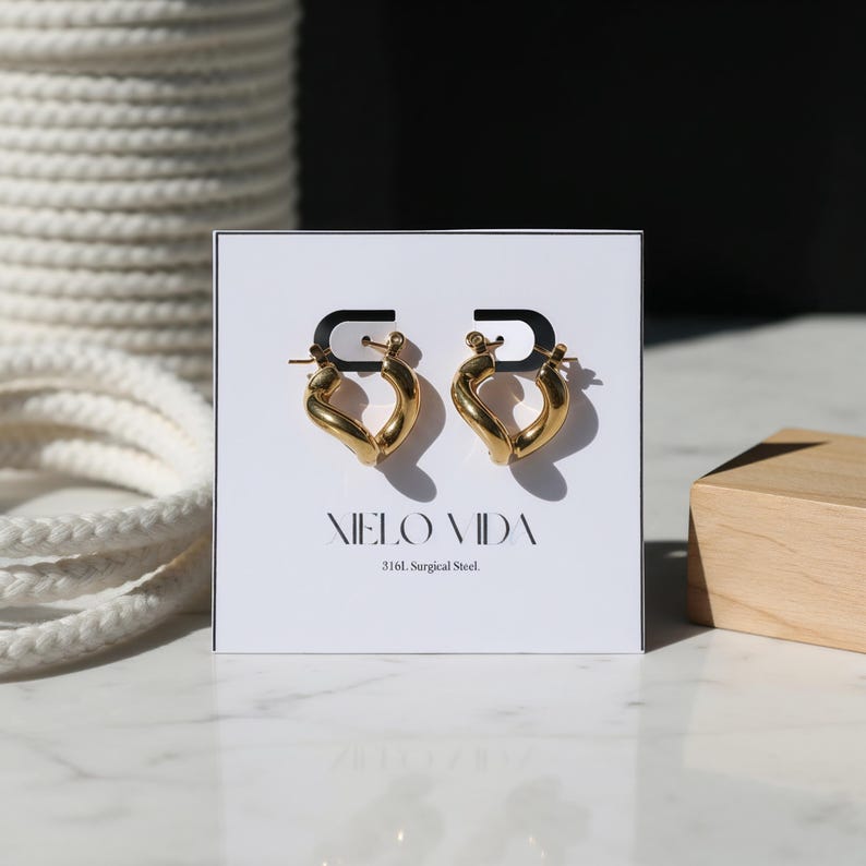 A pair of gold-colored, twisted, hinged hoop earrings are displayed on a white card that reads "XIELO VIDA 316L Surgical Steel" against a dark background with white rope.
