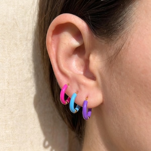 May include: Three small hoop earrings worn in a row on an earlobe. Each hoop features a smooth, vibrant enamel coating in a distinct solid color: one bright hot pink, one sky blue, and one purple. The base of each hoop is a metallic gold tone.