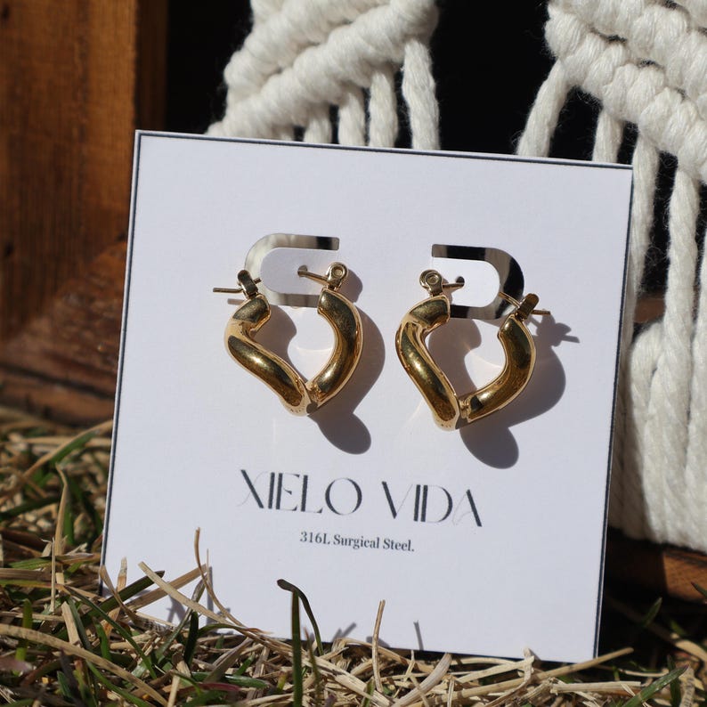 A pair of gold-colored, twisted, hinged hoop earrings are on a white display card in dry grass, with a white macrame hanging in the background.