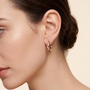 A woman with dark hair wears a gold hoop earring with pink, green, and blue enamel floral designs on her right ear.
