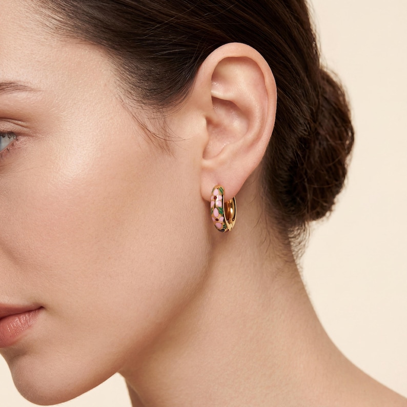 A woman with dark hair wears a gold hoop earring with pink, green, and blue enamel floral designs on her right ear.