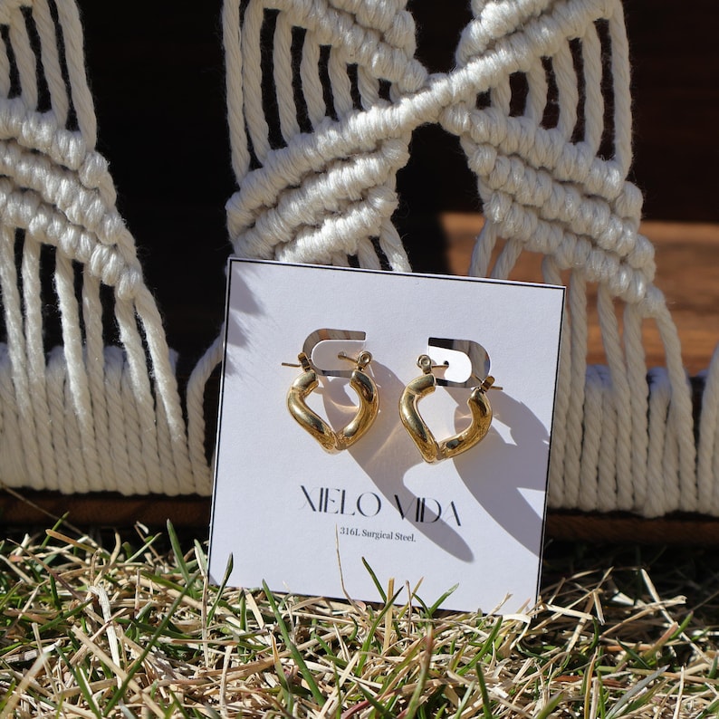 A pair of gold-colored, twisted, hinged hoop earrings are on a white display card in dry grass, with a white macrame hanging in the background.