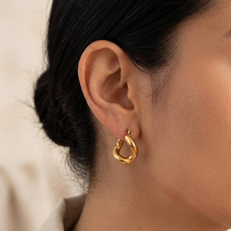May include: A close-up of a gold-colored hoop earring. The earring has a thick, rounded design and is worn on the ear. The earring is shiny and reflects light. The background is blurred, focusing attention on the jewelry.
