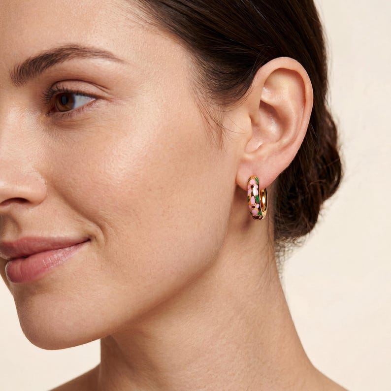 A woman with dark hair and a light-colored top wears a gold hoop earring with pink, green, and blue enamel floral designs on her right ear.