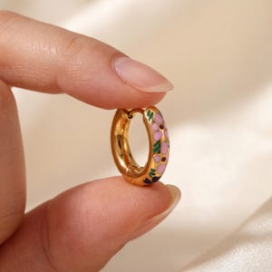 A hand holds a single gold hoop earring with pink, green, and blue enamel floral designs.