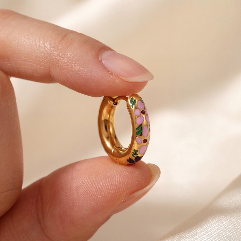 A hand holds a single gold hoop earring with pink, green, and blue enamel floral designs.