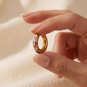 A hand with manicured nails holds a single gold hoop earring with pink, green, and blue enamel floral designs.