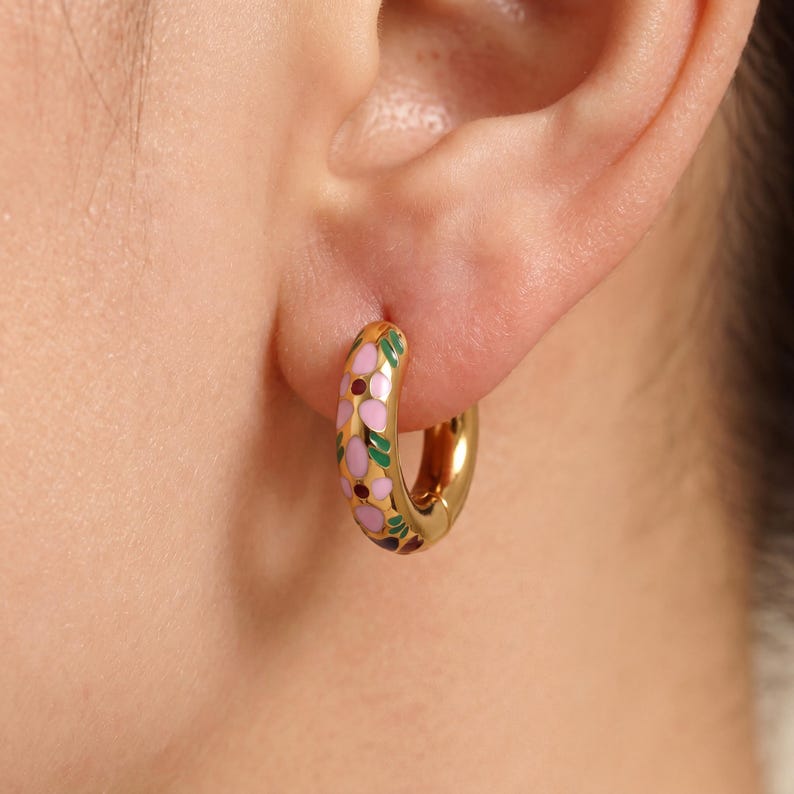 A close-up of a gold hoop earring with pink, green, and blue enamel floral designs worn on an ear.