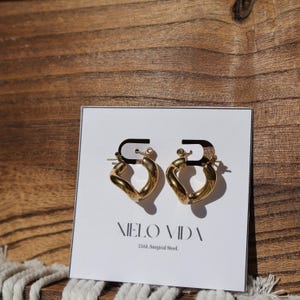 A pair of gold-colored, twisted, hinged hoop earrings are on a white display card in front of a wooden background.