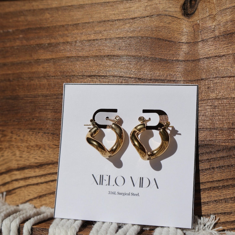 A pair of gold-colored, twisted, hinged hoop earrings are on a white display card in front of a wooden background.
