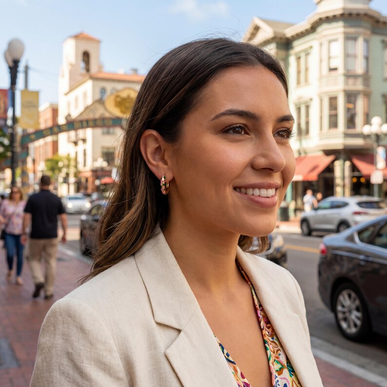 A woman with dark hair smiles, looking right, wearing a cream blazer and a gold floral hoop earring on her right ear, with a city street in the background.