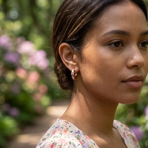 A woman with dark hair in a floral dress looks right, wearing a gold floral hoop earring, with blurred green foliage and pink flowers in the background.