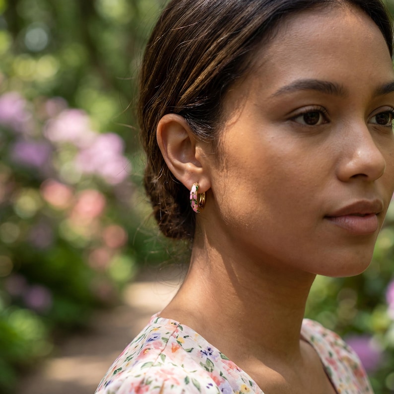 A woman with dark hair in a floral dress looks right, wearing a gold floral hoop earring, with blurred green foliage and pink flowers in the background.