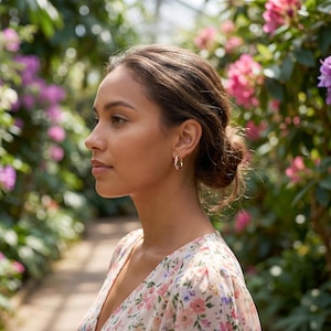A woman with dark hair in a floral dress, looking left, wears a gold floral hoop earring, with blurred pink flowers and greenery in the background.