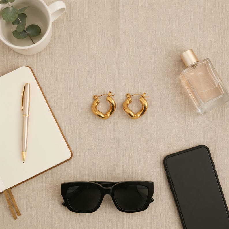 Two gold-colored, twisted, hinged hoop earrings are centered on a light tan fabric surface, surrounded by a pen, notebook, sunglasses, smartphone, and perfume bottle.