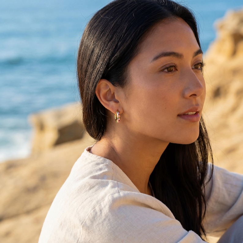 A woman with dark hair wears a gold hoop earring with pink, green, and blue enamel floral designs while looking out at a beach.