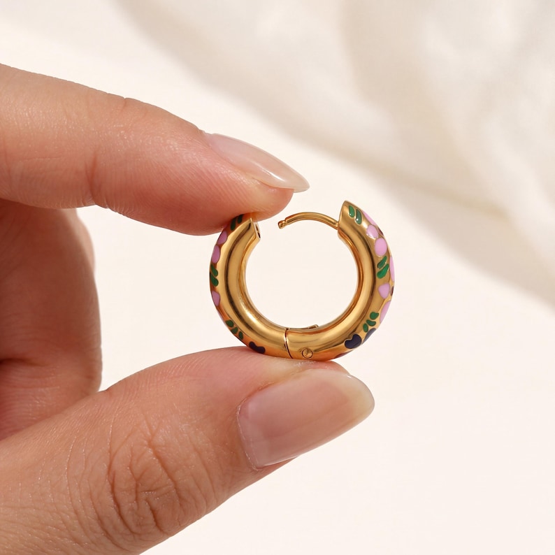 A hand holds a single gold hoop earring with pink, green, and blue enamel floral designs, showing the clasp.