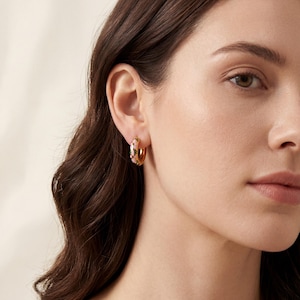 A woman with dark hair wears a gold hoop earring with pink, green, and blue enamel floral designs on her left ear.