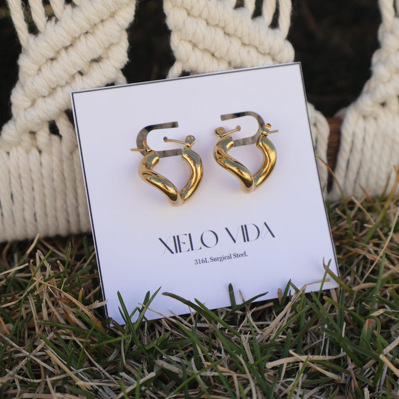 A pair of gold-colored, twisted, hinged hoop earrings are on a white display card in green grass, with a white macrame hanging in the background.