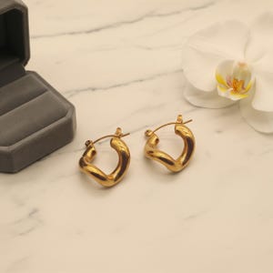 Two gold-colored, twisted, hinged hoop earrings are on a white marble surface next to a gray velvet box and a white orchid.