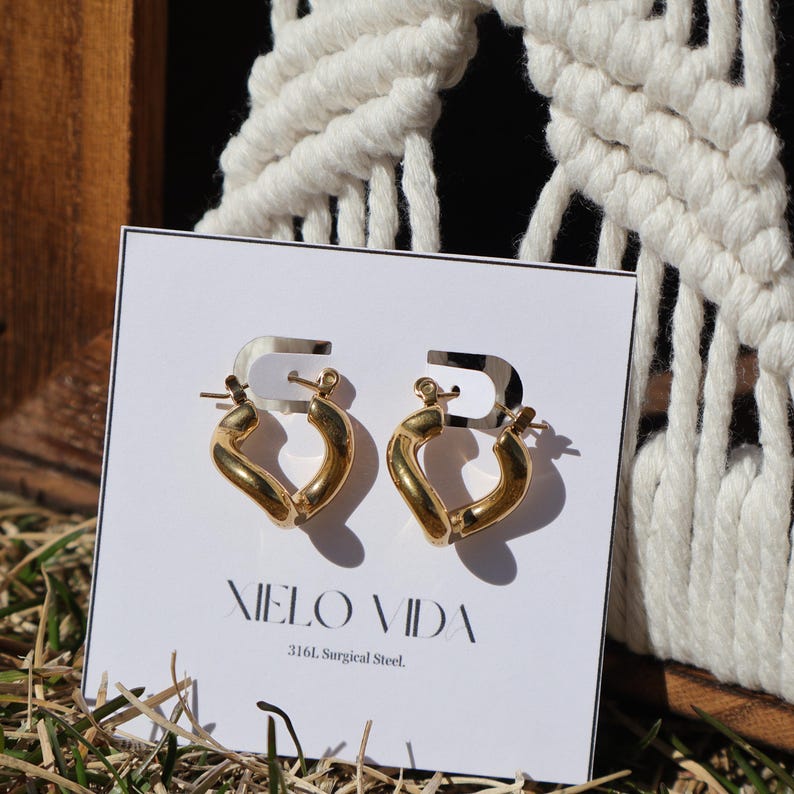 A pair of gold-colored, twisted, hinged hoop earrings are on a white display card in dry grass, with a white macrame hanging in the background.