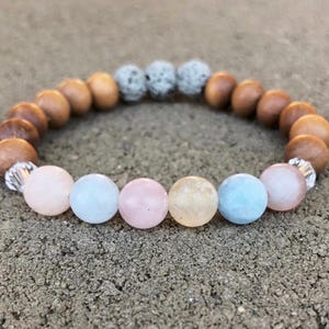 May include: A bracelet with a mix of wooden beads, light blue, pink, and yellow gemstones, and grey lava stone beads. Silver-coloured spacers are also included. The bracelet is displayed on a textured surface.