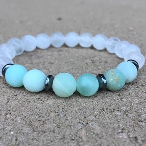 May include: A stretch bracelet with white and turquoise beads. The bracelet incorporates small, dark beads between the larger stones. The bracelet is presented on a textured, neutral-toned surface. The bracelet's beads are measured in centimetres.
