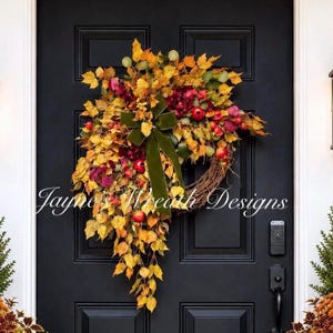 May include: A decorative autumn wreath featuring yellow and orange maple leaves, deep red berries, and small red apples, accented with a large olive green velvet ribbon bow. The wreath is mounted on a black door with the text Jayne's Wreath Designs.